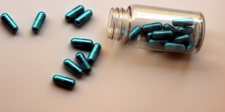 a glass bottle tipped on its side with shiny teal capsules spilling out and scattered on a light surface. uk