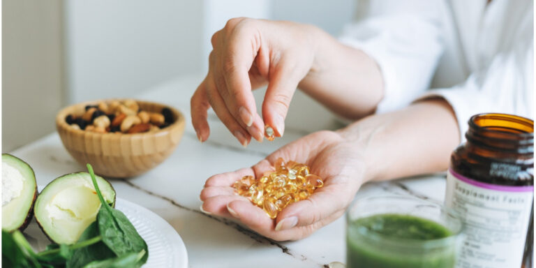 a person holds several yellow softgel capsules in one hand while picking one up with the other. on the table are a bowl of mixed nuts, half an avocado, spinach, a green drink, and a supplement bottle. uk