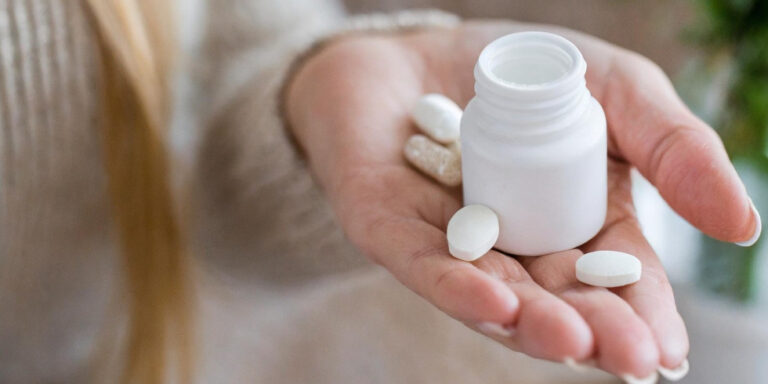a person holds a white pill bottle and several white tablets in their hand, with a few pills resting on their palm. the background is softly blurred. uk