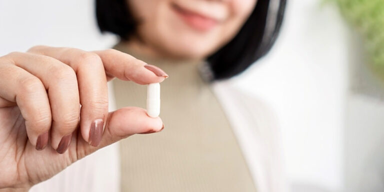 a woman with short dark hair holds a white capsule between her thumb and index finger, smiling softly. the background is softly blurred. uk