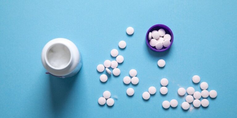 white round tablets scattered on a blue surface, with some inside a purple bottle cap and an open white bottle lying on its side. uk
