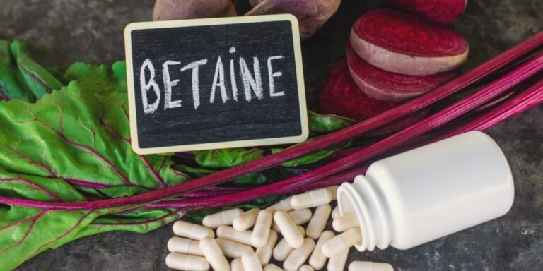 a small blackboard with betaine written on it sits next to fresh beetroot and greens. in front, a white bottle is tipped over with white capsules spilling out onto a dark surface. uk