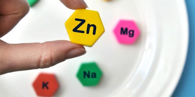 a hand holds a yellow hexagonal tile labelled zn. nearby on a white surface are hexagonal tiles labelled na (green), k (red), and mg (pink), representing chemical elements. uk