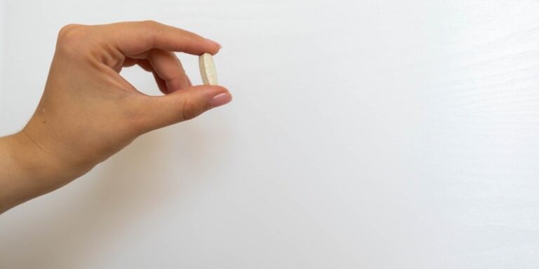 a hand holds a single oval shaped pill or tablet between the thumb and index finger against a plain white background. uk