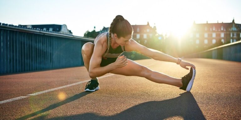 a woman in sportswear stretches her legs on an outdoor track at sunrise or sunset, reaching towards her outstretched foot with one hand while balancing in a deep side lunge. buildings and a bright sun are in the background. uk