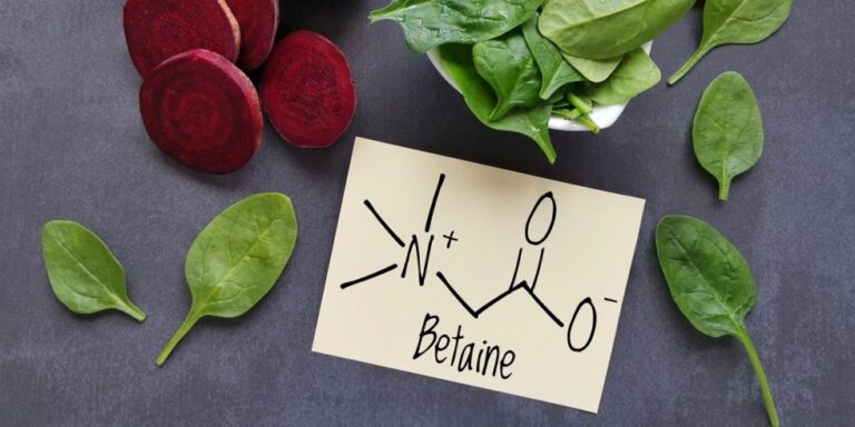 a card with the chemical structure and name “betaine” lies on a dark surface, surrounded by fresh spinach leaves and sliced red beetroot. uk