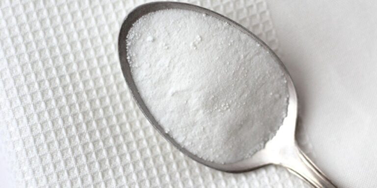 a metal spoon filled with a fine white powder, likely bicarbonate of soda, rests on a textured white surface. uk