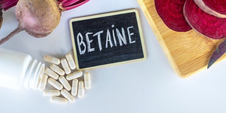 a small blackboard with betaine written on it is surrounded by white capsules, whole and sliced beetroot, and a white bottle on a light surface with a wooden chopping board. uk