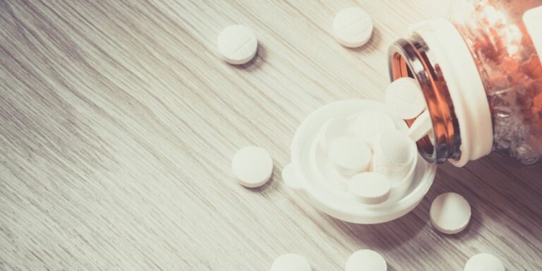 a brown pill bottle tipped over on a light wooden surface, with round white tablets spilling out onto the surface and into the open bottle cap. uk