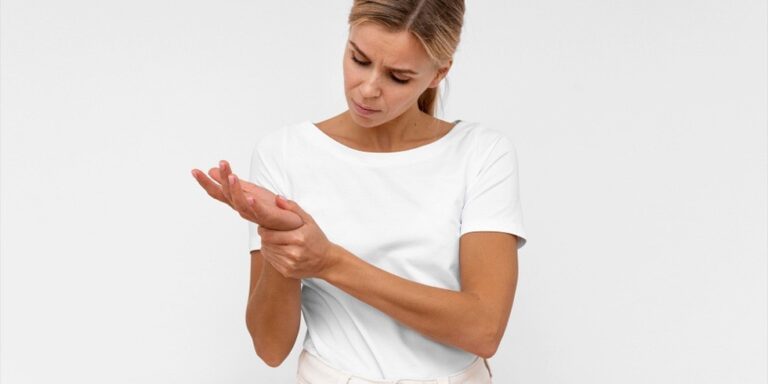 a woman wearing a white t shirt is looking down at her left hand and holding her wrist with her right hand, appearing to be in discomfort or pain. the background is plain and white. uk