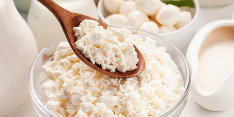 a wooden spoon scoops cottage cheese from a clear glass bowl. other dairy products like mozzarella balls and milk are visible in the background. uk