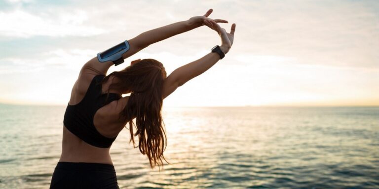 a woman in sportswear stretches her arms overhead by the sea at sunrise or sunset, with her back to the camera. she is wearing a fitness tracker and an armband, and the sky is softly lit. uk