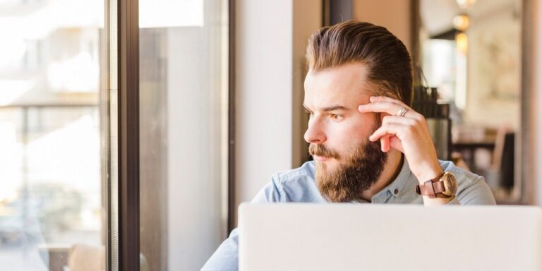 a bearded man with slicked back hair sits at a desk with a laptop, looking thoughtfully out of a window. he rests his hand against his temple and wears a light blue shirt and a watch. uk