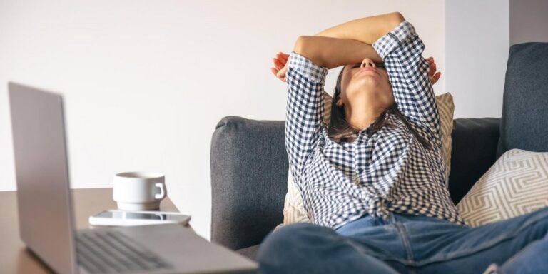 a woman in a chequered shirt leans back on a sofa with her arms over her face, looking tired or stressed. a laptop, mobile phone, and a cup are on the table in front of her. uk