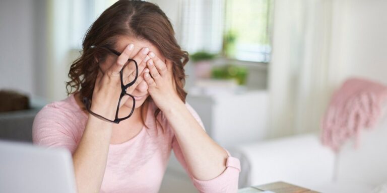 a woman sitting at a desk holds her glasses and covers her face with her hands, appearing stressed or tired. a laptop and papers are on the desk in front of her. uk