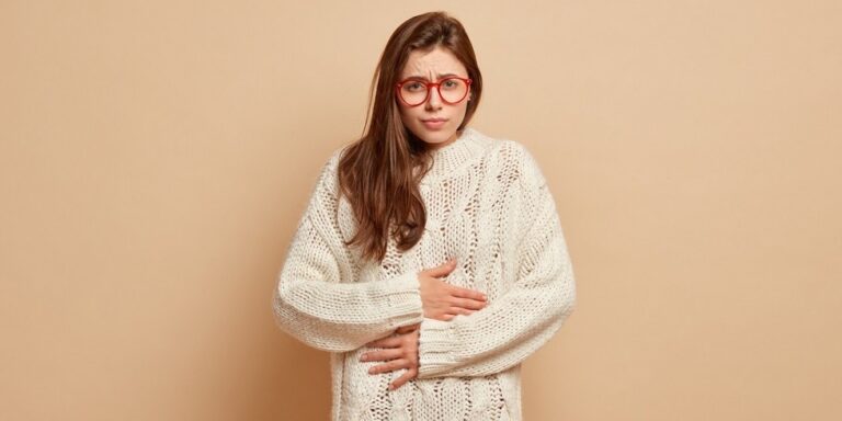 a young woman with long brown hair and red glasses wearing a white knitted jumper holds her stomach with a pained expression, standing in front of a plain beige background. uk
