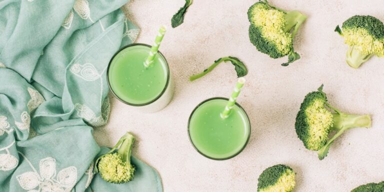 two glasses of green smoothie with striped straws sit on a light surface, surrounded by broccoli florets and a green patterned cloth. uk