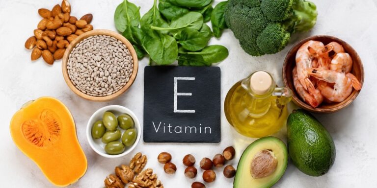 a variety of vitamin e rich foods including prawns, broccoli, spinach, seeds, nuts, avocado, olives, oil, and butternut squash are arranged around a chalkboard with “e vitamin” written on it. uk