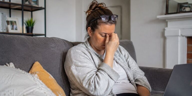 a woman sits on a sofa, eyes closed, pinching the bridge of her nose in apparent frustration or fatigue. she has glasses resting on her head and a laptop in front of her. uk