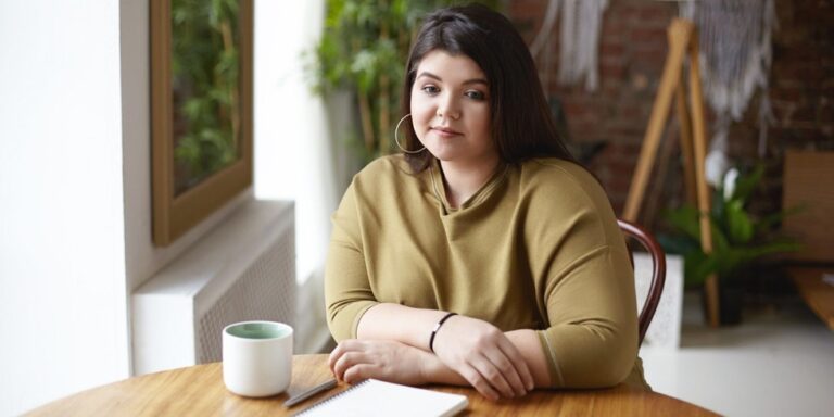 a young woman with long dark hair sits at a wooden table with a notebook, pen, and a white mug. she wears a green top and hoop earrings, looking thoughtfully at the camera in a bright, cosy room with plants. uk