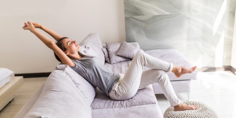 a woman in casual clothes relaxes on a light coloured sofa with her arms stretched above her head and her legs crossed, smiling peacefully in a bright, modern living room. uk