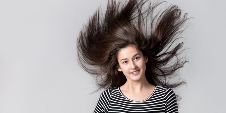 teen girl with long brown hair wearing a black and white striped shirt, smiling at the camera as her hair fans out above her head against a plain, light grey background. uk
